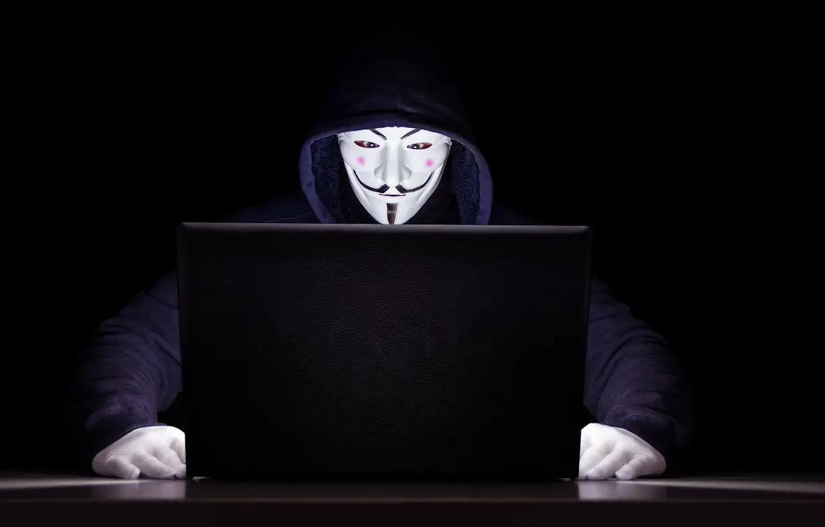 A man wearing an Anonymous V for Vendetta mask sitting behind a laptop, shrouded in darkness.
